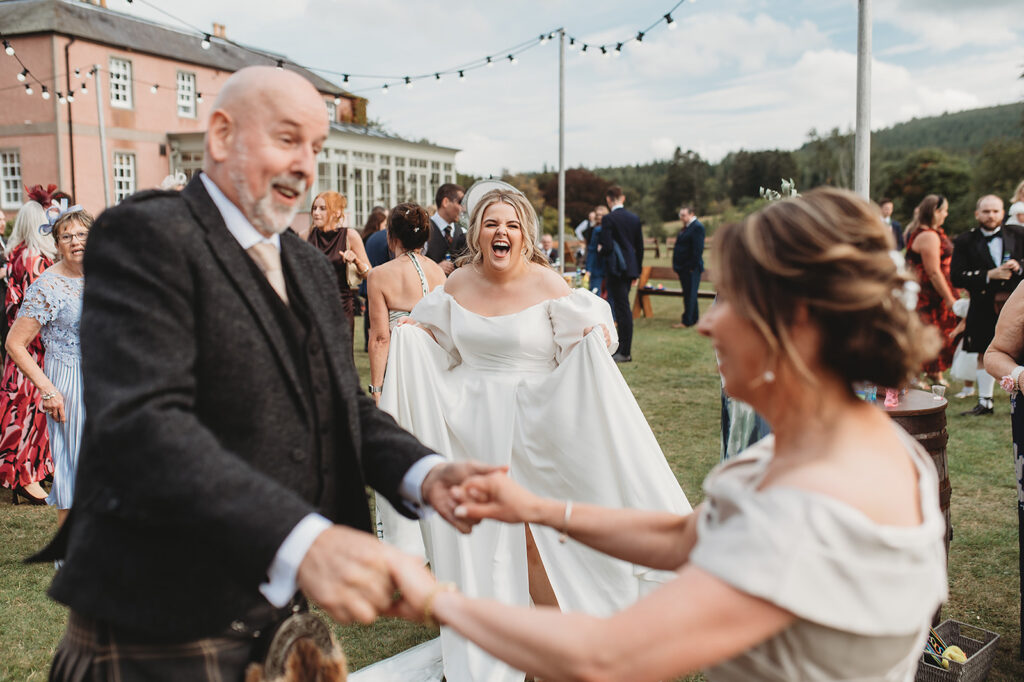 A bride laughing at Ballogie House, Aberdeenshire, with pure joy as she dances with parents on the lawn during her outdoor wedding reception, with a grand pink country house and string lights in the background - captured by Aberdeenshire wedding photographer Samantha Clyne